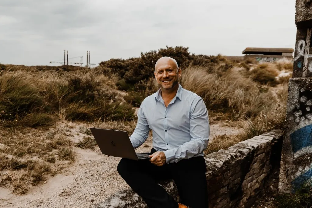Patrick Scholten lachend met zijn laptop op een muurtje in de duinen